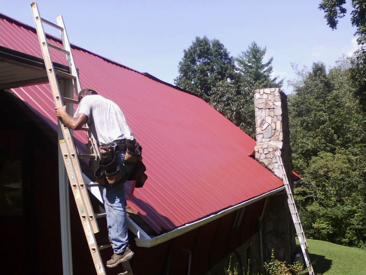 Roofer on a ladder beside a red metal roof and stone chimney — Soffit & Fascia Repair by Roofing Brothers in West New York